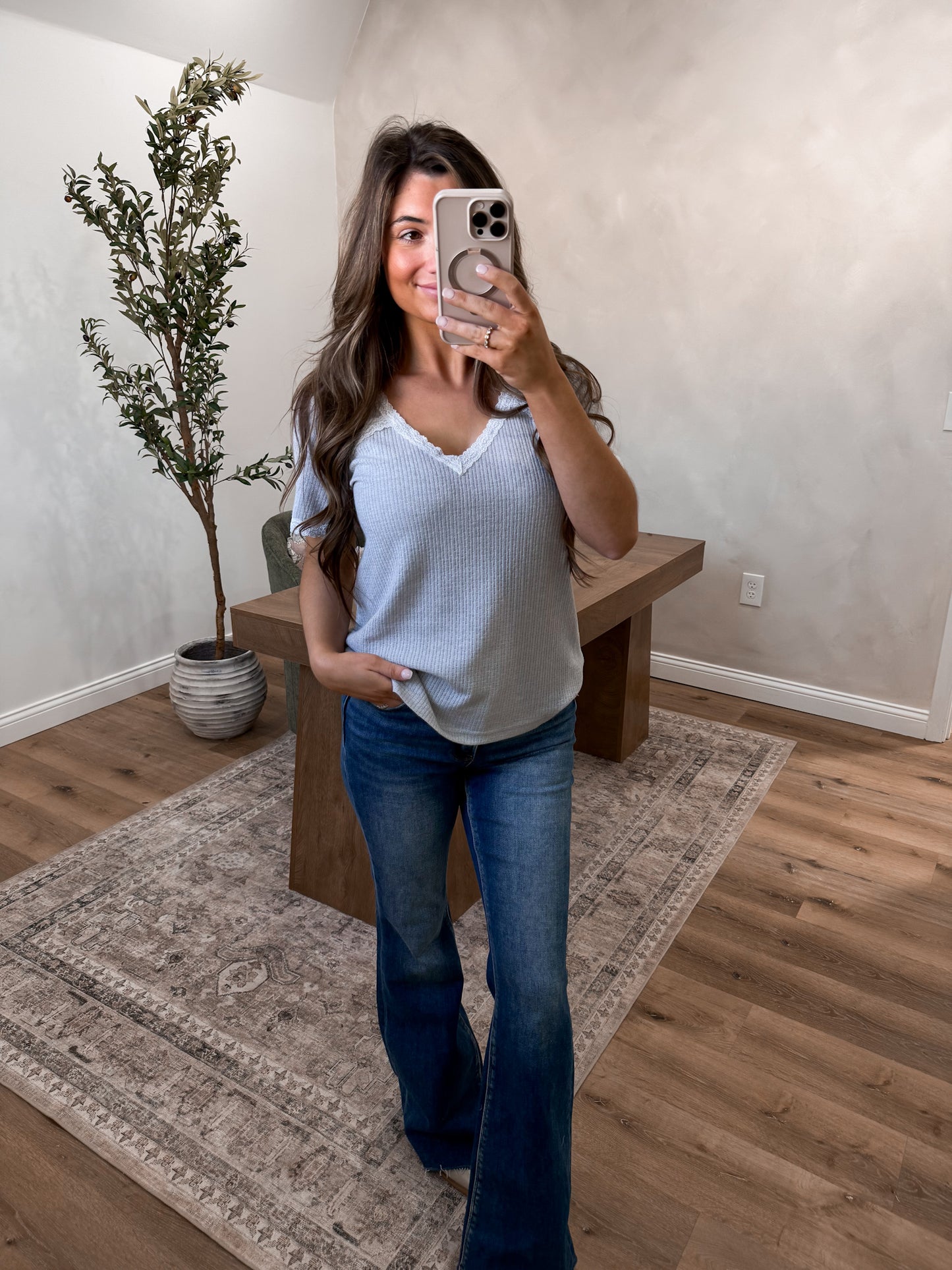 Woman taking a mirror selfie in a room with a plant and wooden bench. Featuring a vneck tee