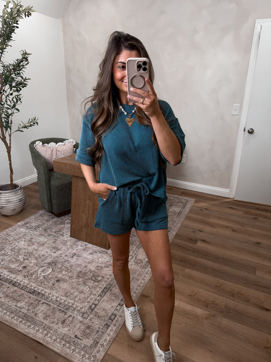 Woman taking a mirror selfie wearing a teal outfit in a room with a plant and chair.