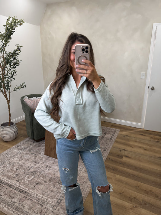 Person taking a mirror selfie wearing a light gray sweater and ripped jeans in a room with a plant and chair.