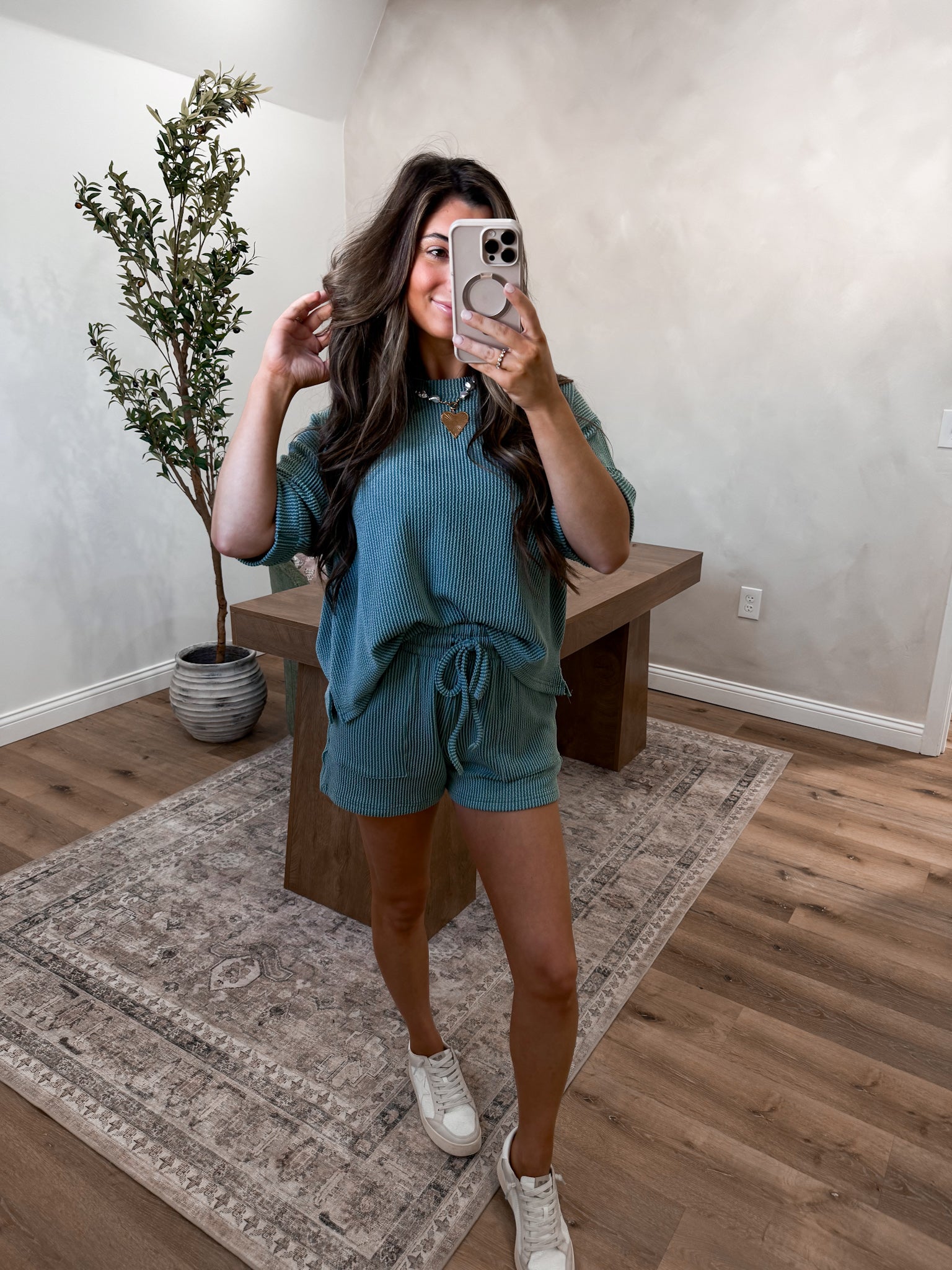 Woman taking a mirror selfie in a teal outfit in a room with a plant and wooden bench.
