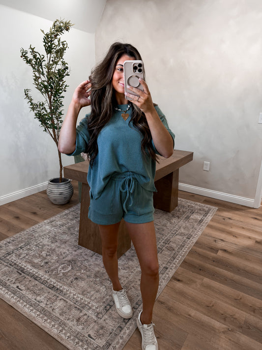 Woman taking a mirror selfie in a teal outfit in a room with a plant and wooden bench.