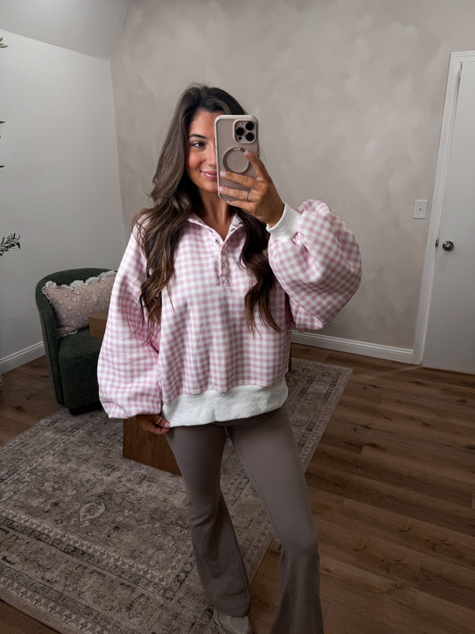 Woman taking a mirror selfie wearing a pink checkered shirt in a room with wooden flooring and a rug.