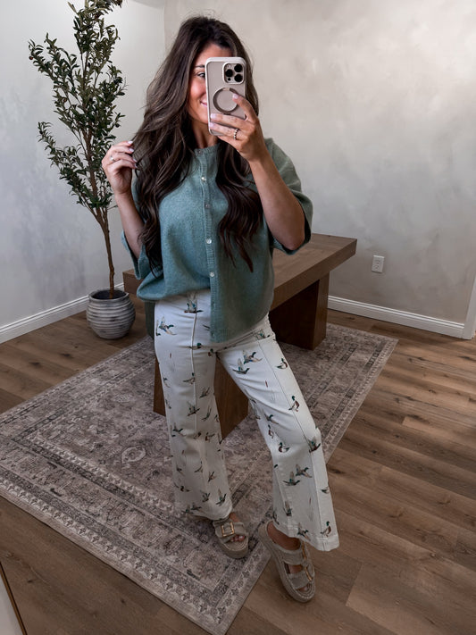 Person taking a mirror selfie wearing a green shirt and patterned pants in a room with a plant and wooden bench.