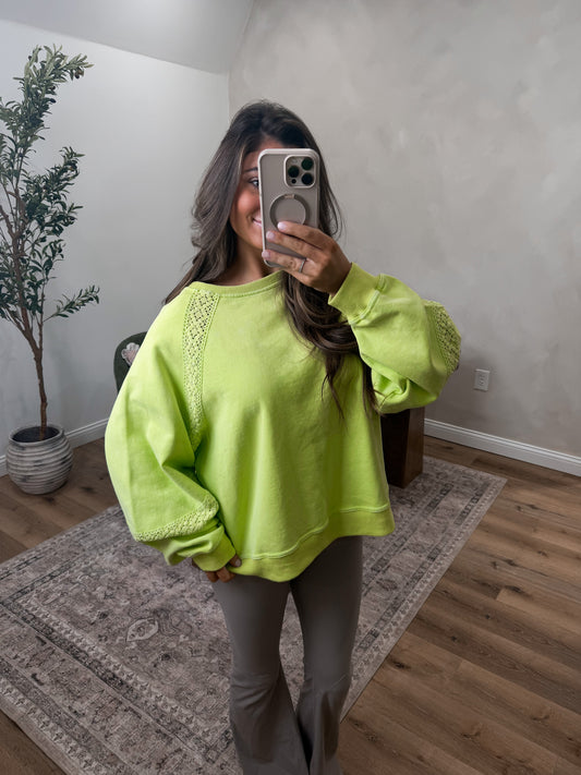 Person wearing a bright green sweatshirt taking a mirror selfie in a room with a plant and wooden floor.