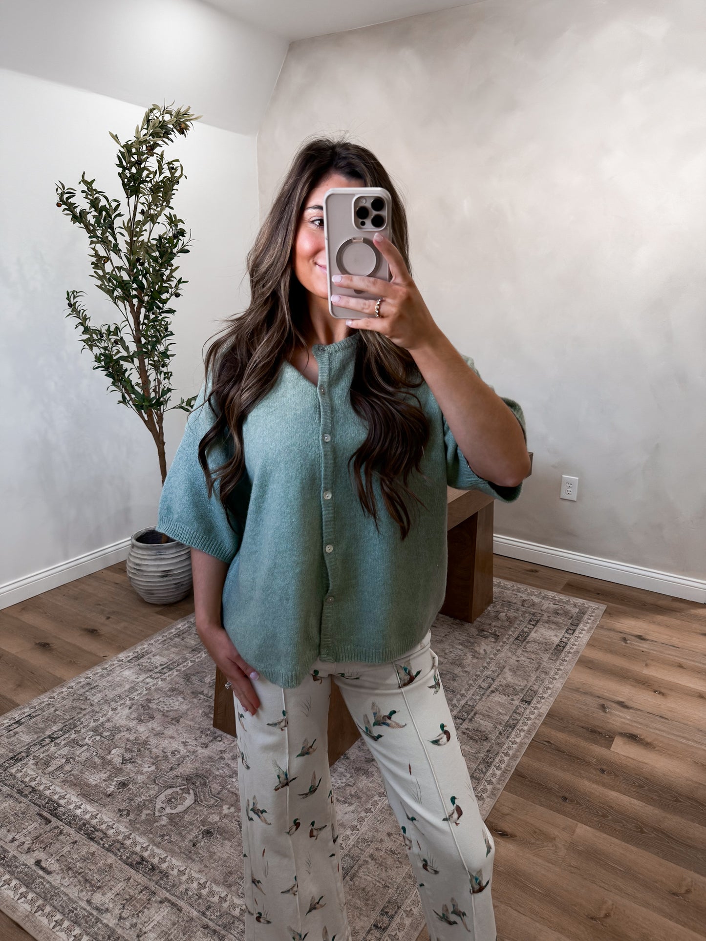 Person taking a mirror selfie wearing a green top and white pants with a pattern, in a room with a plant and wooden floor.