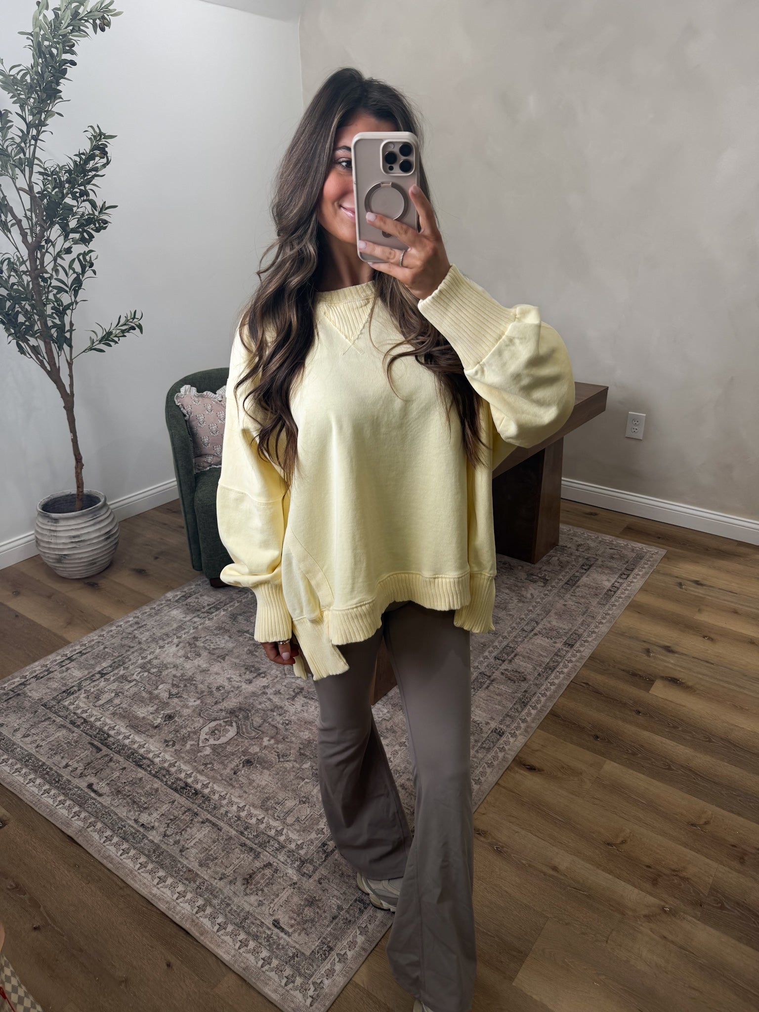 Woman taking a mirror selfie wearing a light yellow sweater and gray pants in a room with a plant and wooden floor.