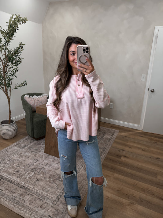 Person taking a mirror selfie wearing a pink sweater and ripped jeans in a room with a plant and chair.