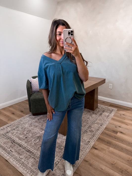 Woman taking a mirror selfie wearing a blue top and jeans in a room with a wooden floor and white walls.