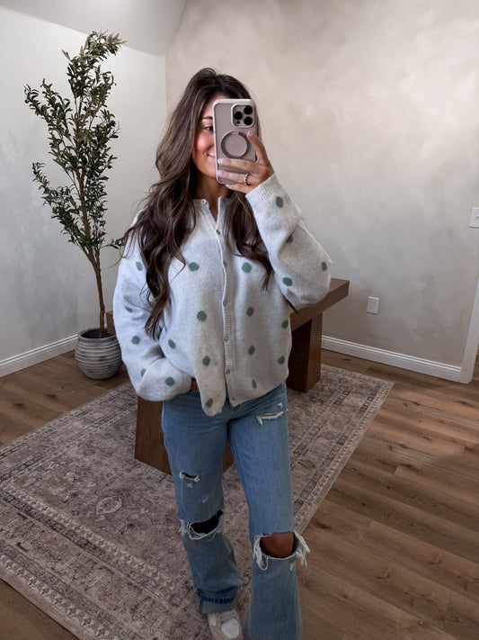 Person wearing a white sweater with green patterns and ripped jeans in a room with a plant and wooden bench.