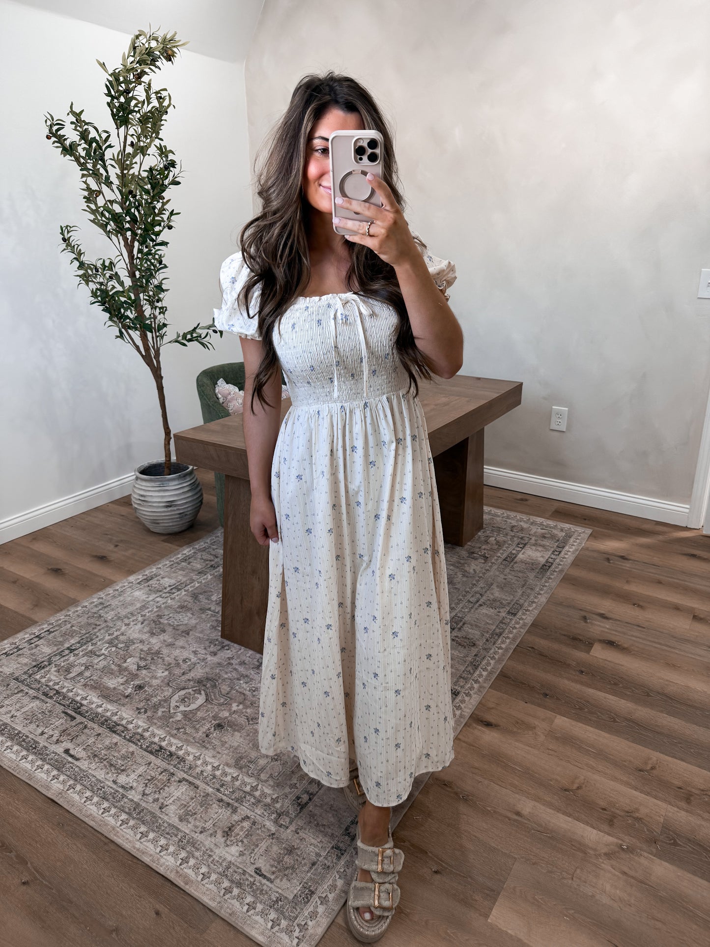 Woman taking a mirror selfie wearing a white dress in a room with wooden flooring and a plant.