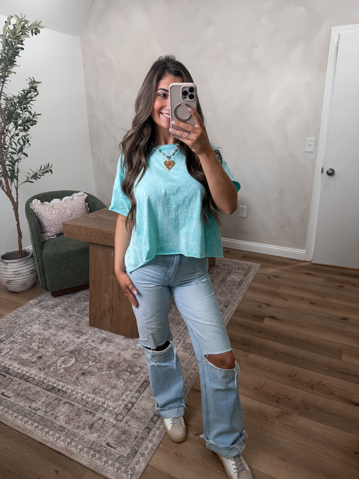 Woman taking a mirror selfie wearing a turquoise top and ripped jeans in a living room.