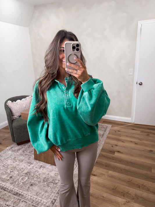 Woman taking a mirror selfie wearing a teal shirt and beige pants in a room with wooden flooring and a gray armchair.
