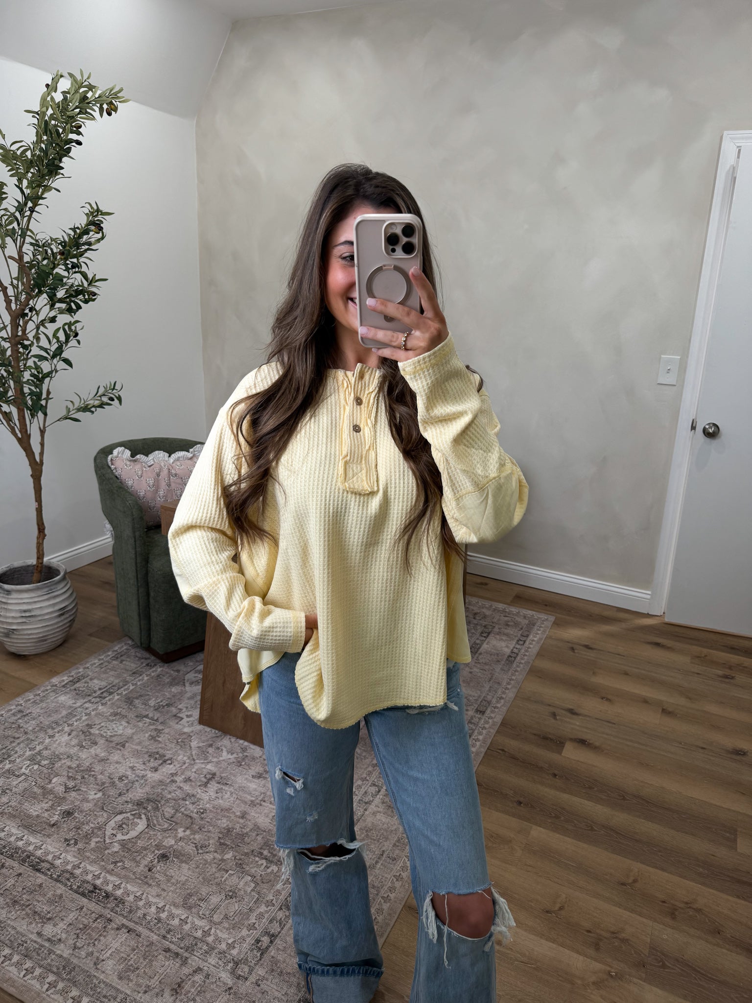 Person wearing a yellow sweater and ripped jeans taking a mirror selfie in a room with a plant and chair.