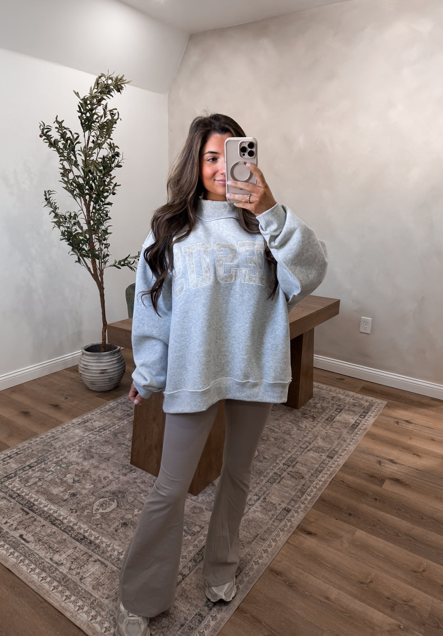 Person taking a mirror selfie wearing a light gray sweatshirt and pants in a room with a plant and wooden bench.