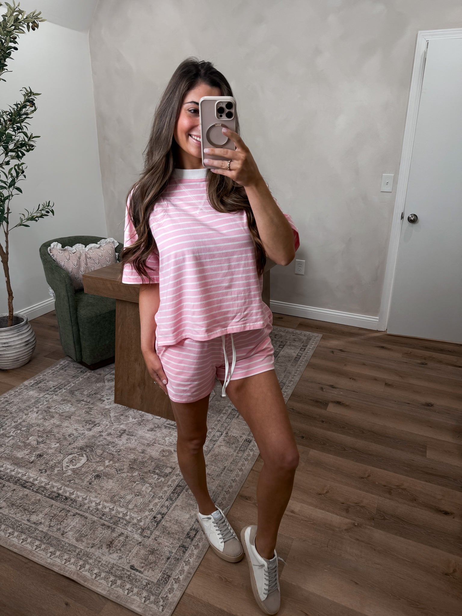Woman taking a mirror selfie wearing a pink striped outfit in a room with a plant and rug.