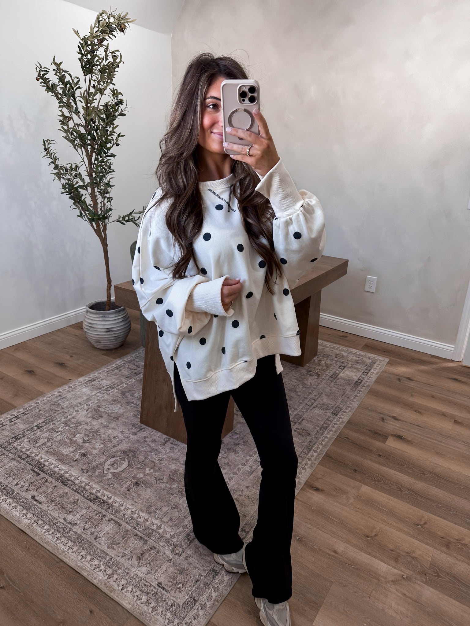 Woman taking a mirror selfie wearing a white sweater with black polka dots and black pants in a room with a plant and wooden bench.