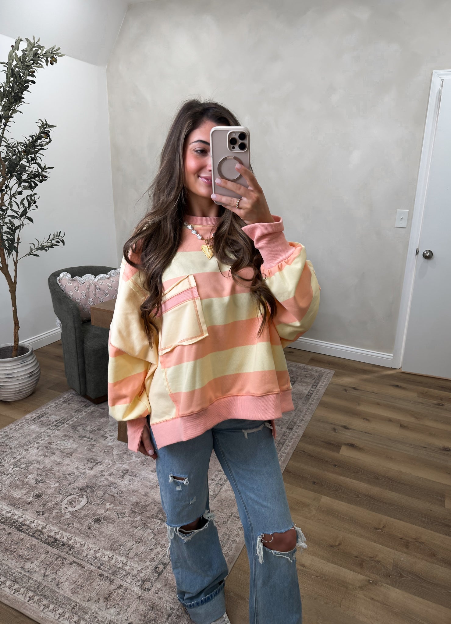 Woman taking a mirror selfie wearing a colorful striped hoodie and ripped jeans in a room with a plant and chair.
