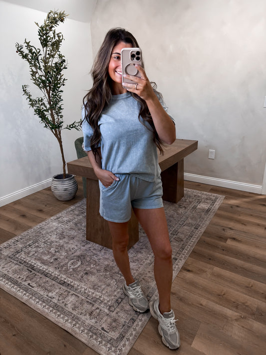 Woman taking a mirror selfie wearing a light blue outfit in a room with a plant and wooden bench.