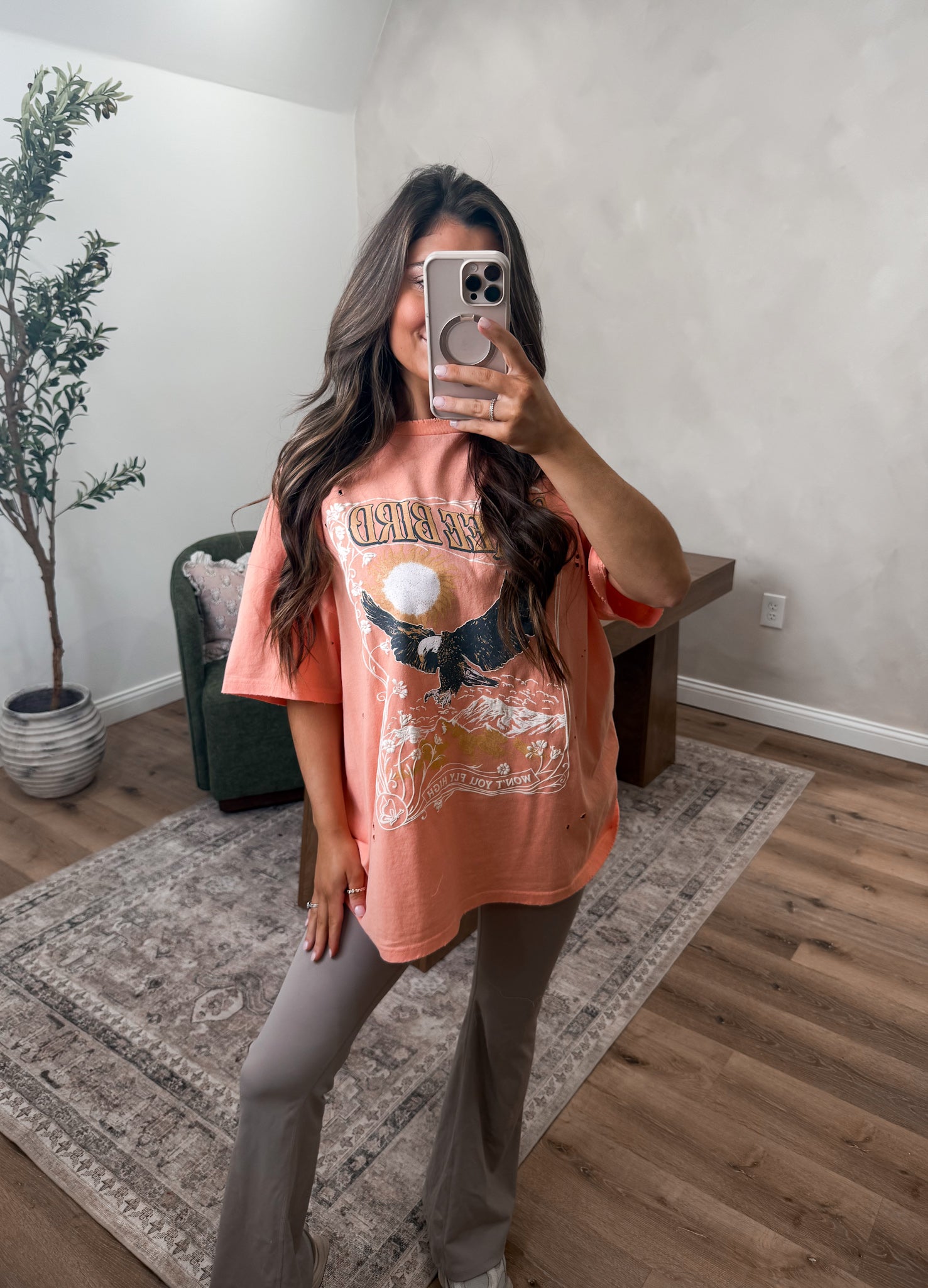 Person wearing a graphic t-shirt and gray pants taking a mirror selfie in a room with a plant and furniture.