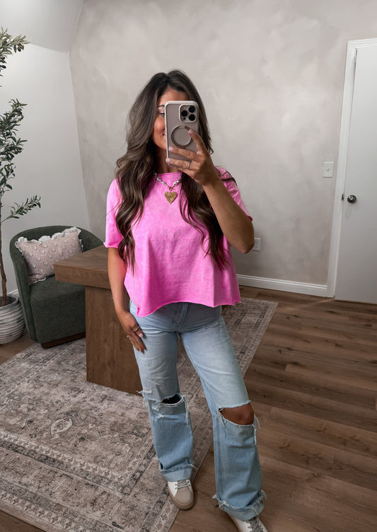Person taking a mirror selfie wearing a pink top and ripped jeans in a room with a wooden floor and white walls.