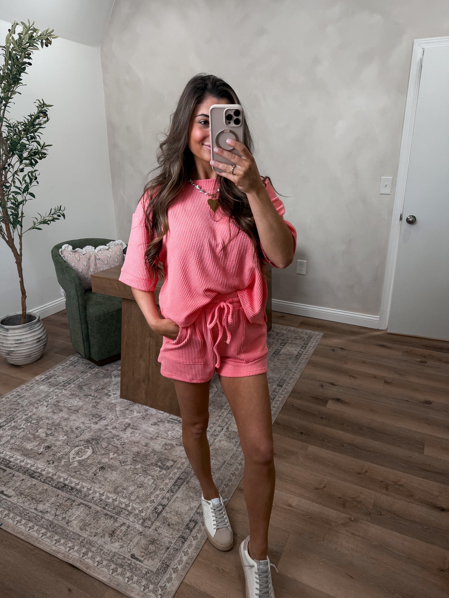 Woman taking a mirror selfie wearing a pink outfit in a room with a plant and rug.