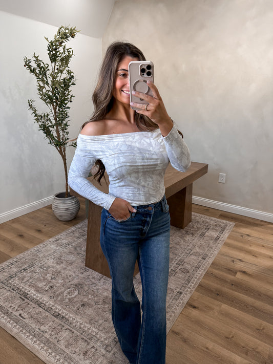 Woman taking a mirror selfie wearing an off-shoulder top and jeans in a room with a plant and wooden bench.