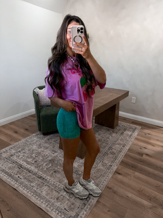 Person taking a mirror selfie wearing a pink floral top and blue shorts in a room with a wooden floor and gray walls.