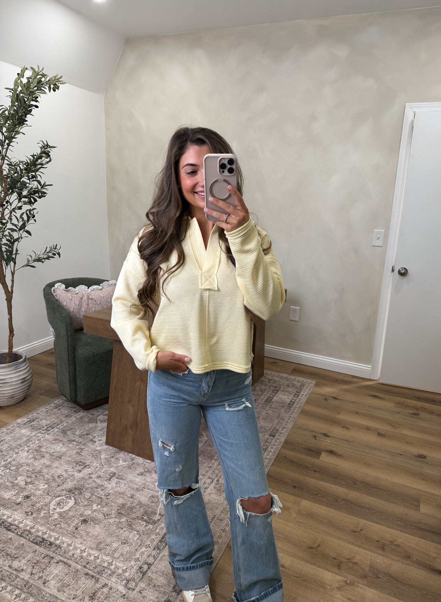 Woman taking a mirror selfie wearing a light yellow hoodie and ripped jeans in a room with a plant and furniture.