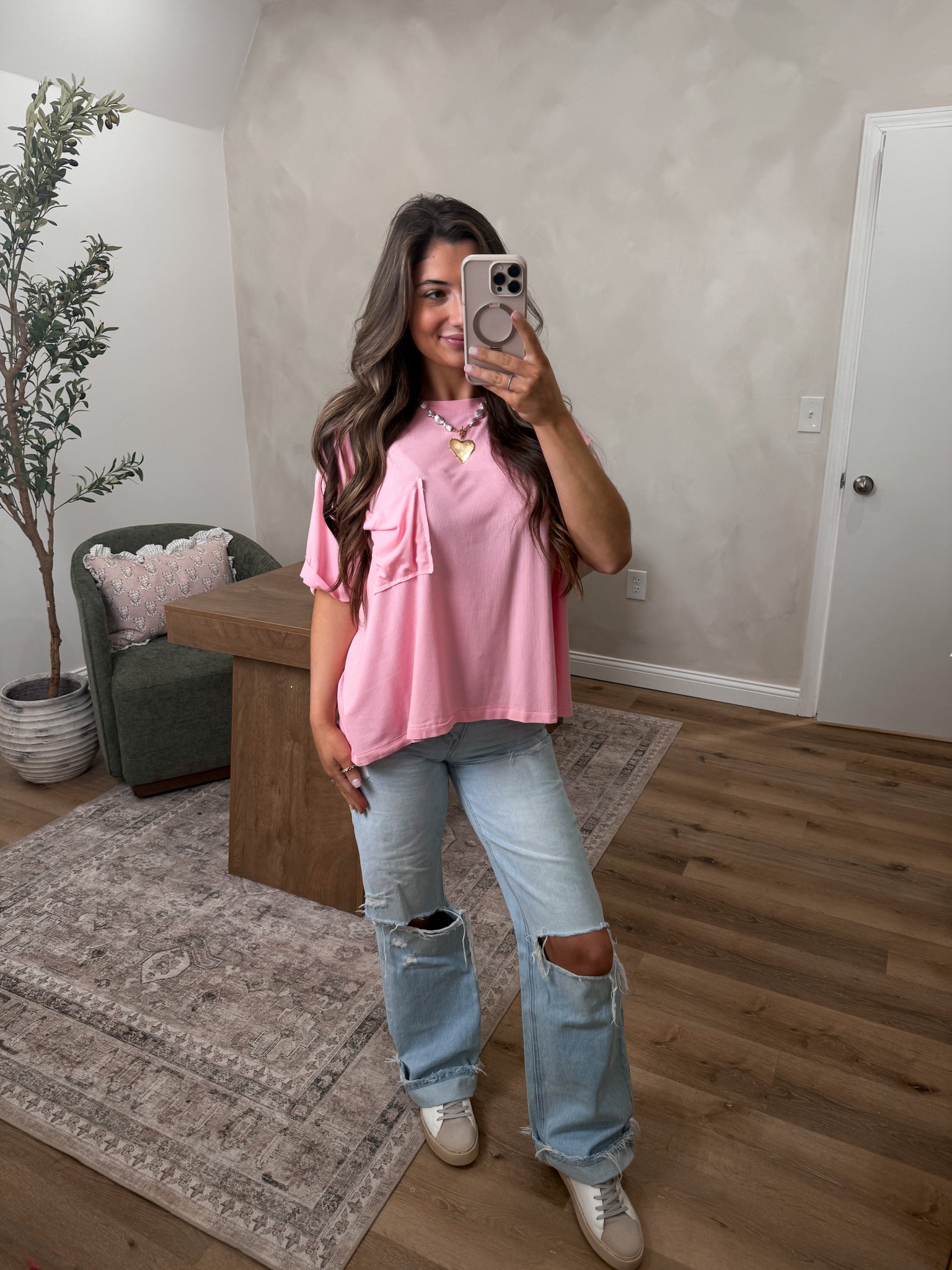 Woman taking a mirror selfie wearing a pink top and ripped jeans in a room with a plant and furniture.