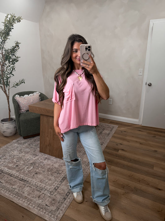 Woman taking a mirror selfie wearing a pink top and ripped jeans in a room with a plant and furniture.