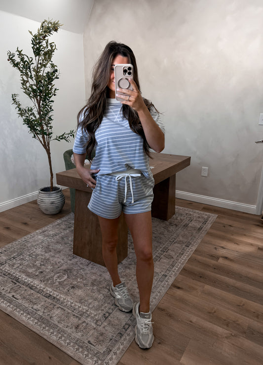 Woman taking a mirror selfie wearing a striped shirt and shorts in a room with a wooden desk and plant.