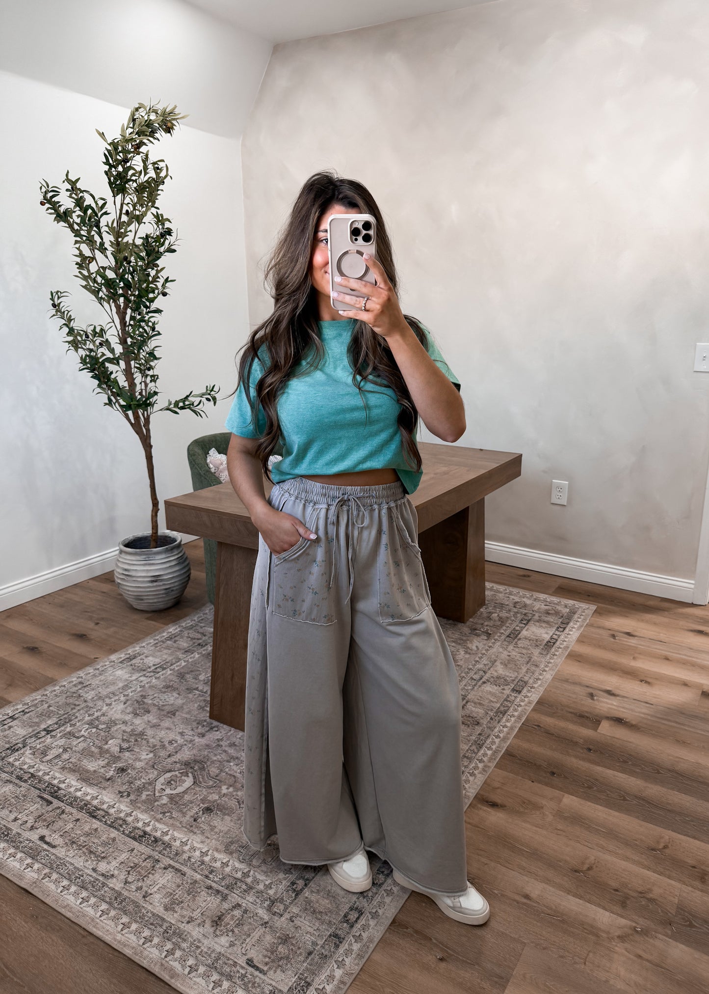 Person taking a mirror selfie wearing a teal top and gray pants in a room with a plant and wooden bench.