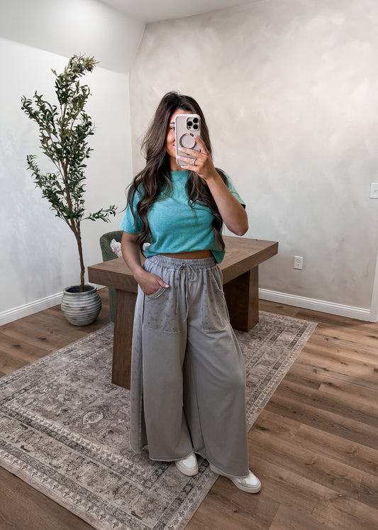 Person taking a mirror selfie wearing a teal top and gray pants in a room with a plant and wooden bench.